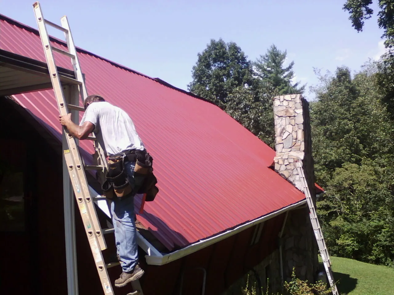Roofer on a ladder beside a red metal roof and stone chimney — Soffit & Fascia Repair by Roofing Brothers in Laredo